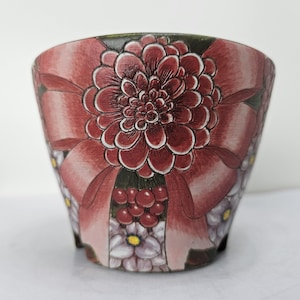 Könnte beinhalten: Ein konischer Keramikübertopf mit einer großen, dunkelroten Dahlie als zentralem Motiv. Das Design umfasst kleinere hellviolette Blüten mit gelben Mitten, rote Beerenbüschel und grünes Blattwerk auf einem dezenten, farblich abgestimmten Hintergrund.
