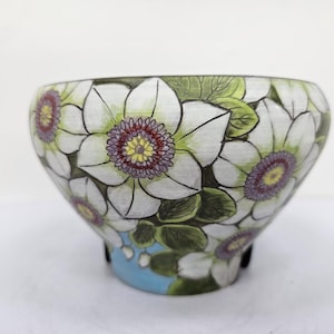 May include: A ceramic bowl featuring a floral pattern. The bowl is adorned with white flowers with purple and yellow centres, green leaves, and a light blue base. A decorative item.