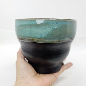 May include: A handcrafted ceramic bowl with a dual-tone glaze. The upper section is teal-green, while the lower part is a dark, near-black hue. The bowl, held in a hand, has a rounded form. The bowl's diameter is approximately 15 cm.