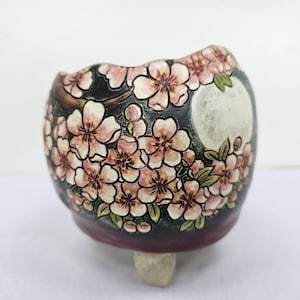 Può includere: Un vaso di ceramica con sfondo scuro, decorato con fiori di ciliegio rosa e bianchi. Il vaso ha una forma arrotondata e un piccolo piede colorato chiaro. I fiori hanno foglie verdi.