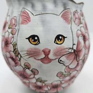 Puede incluir: Jardinera de cerámica pintada a mano con forma de gato. La cara del gato blanco tiene detalles en rosa y grandes ojos amarillos. Flores rosas y una mariposa decoran el exterior de la jardinera. Un artículo decorativo único.
