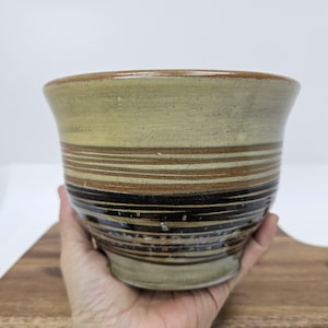 May include: A handcrafted ceramic bowl with a wide, slightly flared rim. The bowl features horizontal bands of varying widths in shades of beige, brown, and black. The bowl is held in a hand, showcasing its size and shape.