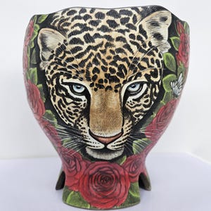 Könnte beinhalten: Eine dekorative Vase mit einem Leopardenkopf, blauen Augen und geflecktem Fell. Die Vase ist mit roten Rosen und grünen Blättern auf schwarzem Hintergrund verziert. Der Sockel hat eine einzigartige Form.