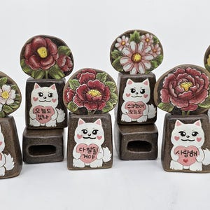 May include: A collection of ceramic cat figurines with floral accents. Each figurine features a white cat holding a heart, with Korean text. The cats are on brown rectangular bases, topped with painted flowers.