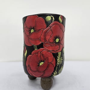 Könnte beinhalten: Schwarzer Keramik-Blumentopf mit geprägten roten Mohnblumenmotiven. Der zylindrische Topf steht auf drei kleinen, abgerundeten Füßen. Die Mohnblumen haben grüne Zentren und sind vor einem schwarzen Hintergrund angeordnet.