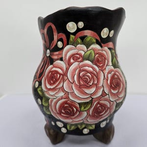 Pode incluir: Um vaso de cerâmica pintado à mão com base preta e um buquê de rosas cor-de-rosa. O vaso tem uma fita decorativa e detalhes de pérolas. O vaso é suportado por três pés pequenos.