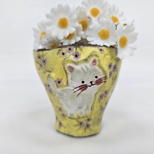 May include: A yellow ceramic vase with a white cat design and purple floral accents, filled with white daisies. The vase is hand-painted and has a unique, artistic appearance, perfect for home decor.