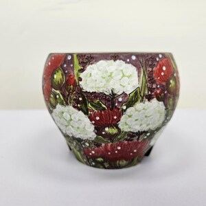 May include: A round, decorative ceramic pot with a floral design. The pot features white hydrangea blooms, red mushroom-like accents, and green foliage against a dark background. The pot is approximately 5 inches in diameter.