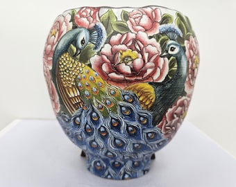 May include: A decorative ceramic vase featuring a hand-painted design of two peacocks amidst pink peonies and green foliage. The vase has a blue base with small feet and a scalloped rim. The colors include blue, yellow, pink, and green.