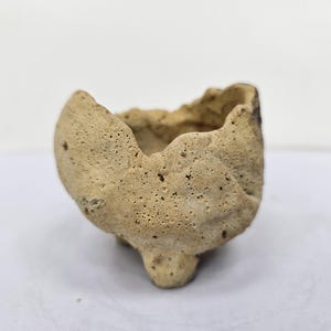 Puede incluir: Pequeño cuenco de cerámica beige hecho a mano, con una superficie rugosa y texturizada. El cuenco tiene una forma irregular con una base redondeada y un pie pequeño. El borde es desigual.