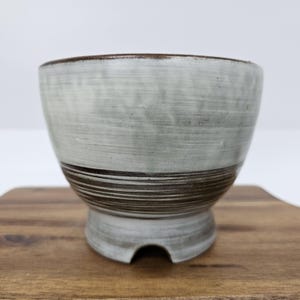 May include: A handmade ceramic bowl with a light gray glaze and a dark brown band around the middle. The bowl has a footed base and a dark brown rim. The bowl is sitting on a wooden surface.
