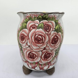 Puede incluir: Jardinera de cerámica con un diseño floral pintado a mano. La jardinera presenta un grupo de rosas rosas con hojas verdes sobre un fondo blanco. El borde es festoneado y la base tiene tres pequeños pies.