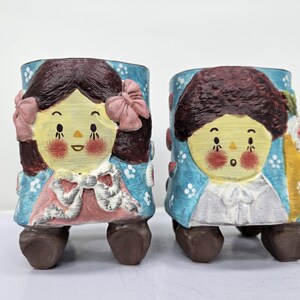May include: Two ceramic planters with a whimsical design. Each planter features a cartoon-style girl with brown hair, rosy cheeks, and a blue dress. The planters are supported by brown feet.