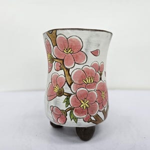 May include: A white ceramic vase with a floral design. The vase features pink blossoms with yellow centers and brown branches. The vase has a slightly curved shape and rests on three small, dark brown feet.