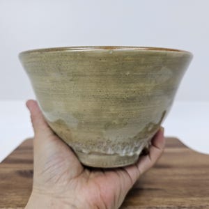 May include: A handmade ceramic bowl with a wide, rounded shape. The bowl has a light green and tan color with a textured surface. The rim has a darker brown hue. The bowl is held in a hand.