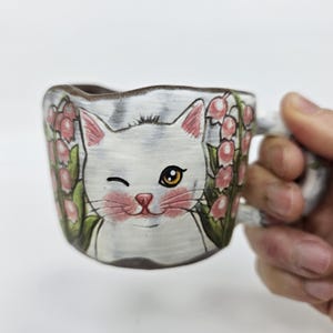 Puede incluir: Taza de cerámica con un gato blanco guiñando un ojo, detalles rosas y diseños florales. La taza tiene un asa texturizada y está sujeta por una mano. El gato tiene un ojo amarillo y mejillas rosas. Fondo blanco.