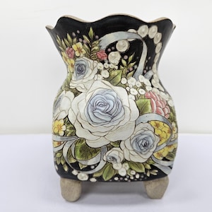 Puede incluir: Un jarrón decorativo de cerámica con fondo negro y diseño floral. El jarrón presenta rosas blancas, azules y rosas, con detalles amarillos y blancos. El jarrón tiene un borde festoneado y se apoya en tres pequeños pies.