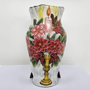 Puede incluir: Un jarrón decorativo de cerámica alto con un diseño festivo. El jarrón presenta una vela, flores de pascua rojas y bayas. El jarrón está pintado con una paleta de colores blanco, gris, amarillo, rojo y verde. El jarrón mide aproximadamente 25 cm de alto.