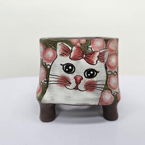 Puede incluir: Una maceta cuadrada de cerámica con un diseño de gato pintado a mano. El gato tiene un lazo rojo, bigotes y mejillas rosadas. La maceta es blanca con detalles florales rosas y patas marrones.