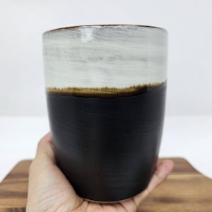 May include: A ceramic tumbler with a two-tone design. The top half is white with brushstrokes, transitioning to a dark brown band, and then a black base. The mug is held in a hand, showing its size and shape. A handmade item.