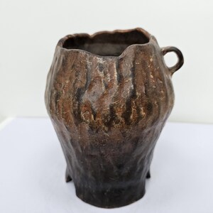 Peut inclure: Un vase en céramique marron fait à la main avec une surface texturée ressemblant à de l'écorce. Le vase a une forme irrégulière, une petite anse et trois pieds. Décoration d'intérieur.