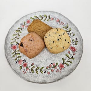Puede incluir: Un plato de cerámica gris con un diseño floral contiene tres galletas. Una es una galleta rectangular marrón claro, otra es una galleta cuadrada rosa y la tercera es una galleta redonda con motas negras. El plato tiene un borde decorativo de flores rosas y hojas verdes.