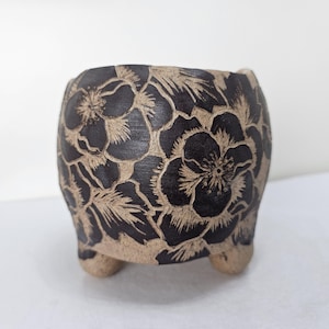 May include: A small, rounded ceramic planter featuring three short feet and a dark brown exterior surface. The pot is decorated with a carved, light beige floral pattern that reveals the natural clay color beneath the dark finish.
