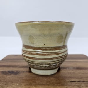 May include: A handmade ceramic cup with a wide, flared rim and a rounded base. The cup features a light beige glaze with horizontal bands of brown and cream-colored stripes. The cup is approximately 4 inches tall and 4 inches wide.