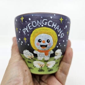Puede incluir: Una taza de cerámica pintada a mano con un diseño caprichoso. La taza presenta un muñeco de nieve con una capucha amarilla y una bufanda naranja, rodeado de ovejas en un campo verde. La palabra "PYEONGCHANG" está escrita sobre el fondo azul oscuro con estrellas.