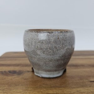 May include: A small, handcrafted ceramic bowl with a speckled gray glaze. The bowl has a rounded shape and sits on three small feet. The interior is a darker brown, and the exterior has a textured appearance. The bowl is suitable for serving small portions.
