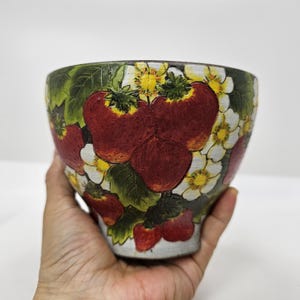 May include: A ceramic bowl featuring a hand-painted design of bright red strawberries, green leaves, and small white flowers with yellow centres. The bowl has a rounded shape and a textured surface, showcasing a vibrant botanical pattern throughout.