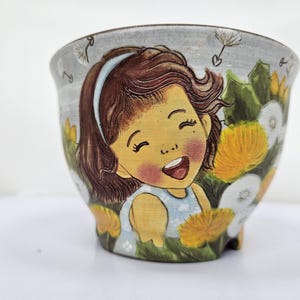 May include: A ceramic planter with a hand-painted illustration of a smiling girl surrounded by dandelions. The planter features a light blue background with yellow and white flowers, and a light blue headband.