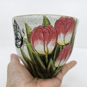 May include: A ceramic bowl with a hand-painted design of red tulips and a black and white butterfly. The bowl has a cream-colored background and green leaves. The bowl is held in a person's hand.