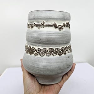 May include: A gray ceramic vase with a textured surface and a decorative floral pattern. The vase has a bulbous shape with two bands of embossed floral designs in a darker brown color.