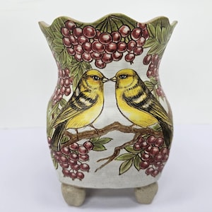 Puede incluir: Un jarrón de cerámica con borde festoneado, con un diseño pintado a mano de dos pájaros amarillos posados en una rama, rodeados de bayas rojas y hojas verdes. El jarrón está sostenido por tres pequeños pies.