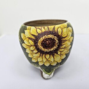 May include: A ceramic pot with a hand-painted sunflower design. The pot features a yellow sunflower with a brown center and green leaves. The pot has a rounded shape and a small, pointed base.