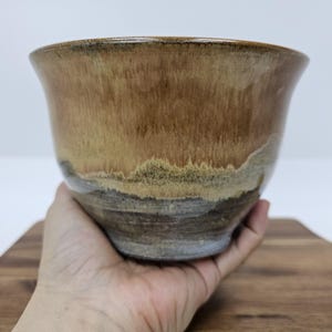May include: A handmade ceramic bowl with a flared rim. The bowl features a gradient of colors, transitioning from a dark brown at the top to lighter tan and gray tones towards the base. The bowl is held in a hand.