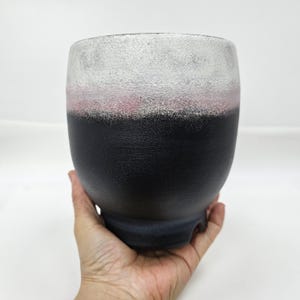 以下が含まれることがあります： ユニークな色のグラデーションを持つ、手作りのセラミック花瓶。花瓶は、テクスチャ加工された黒いベースからピンクのバンド、そしてシルバーホワイトのリムへと変化します。花瓶は丸みを帯びた形状で、3つの小さな足の上に立っています。