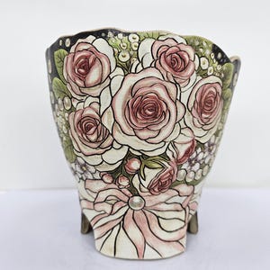 May include: A hand-painted ceramic planter featuring a bouquet of pink roses and white flowers. The planter has a black rim and is decorated with a pink bow and pearl accent. The planter is supported by three small feet.