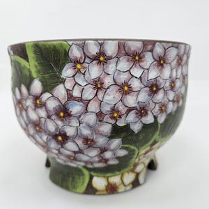 Pode incluir: Uma tigela de cerâmica com um design floral. A tigela apresenta um padrão pintado à mão de flores brancas e roxas com centros amarelos, rodeadas por folhas verdes. A tigela tem uma borda e base marrom escura e está sobre um fundo branco.