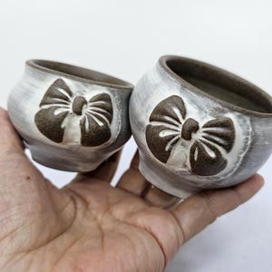 以下が含まれることがあります： 2つの小さな手作りのセラミックボウル。ボウルはグレーと白で、前面に茶色のリボン模様があります。ボウルは人の手で持たれています。
