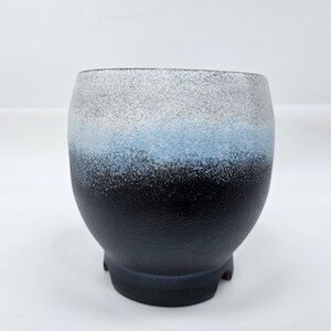 Puede incluir: Una taza de cerámica con un diseño degradado. La parte superior es gris, pasando a azul y luego a negro en la base. La taza tiene forma redondeada y se apoya en tres pequeños pies.