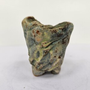 Puede incluir: Una pequeña maceta de cerámica texturizada con una forma orgánica única. La maceta presenta una mezcla de tonos verdes, azules y marrones apagados, con una superficie rugosa e irregular. Tiene una pequeña base de tres patas y una parte superior abierta.