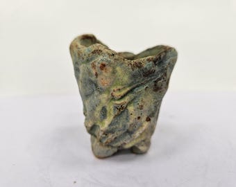 Peut inclure: Petit pot en céramique texturé de forme organique. Le pot présente un mélange de teintes vertes, bleues et brunes, avec une surface rugueuse et inégale. Il possède une petite base à trois pieds et un dessus ouvert.