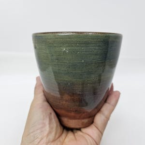 Puede incluir: Una taza de cerámica hecha a mano con un degradado de color único. La mitad superior es verde oscuro, que pasa a un marrón rojizo en la base. El borde es de un marrón más oscuro. La taza se sostiene en una mano sobre un fondo blanco.