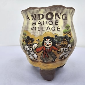 Puede incluir: Una maceta de cerámica en tonos beige y marrón, decorada con una escena pintada a mano de figuras con vestimenta tradicional coreana. El texto en la maceta dice "ANDONG HAHOE VILLAGE".