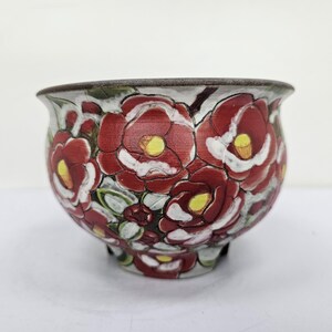 以下が含まれることがあります： 花柄の陶器鉢。 赤い花に黄色の中心と白いアクセントが描かれています。 リムは濃い茶色で、ベースには3つの小さな足が付いています。