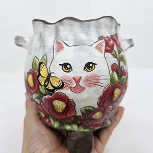 Può includere: Vaso in ceramica bianca con un disegno a forma di gatto, circondato da fiori rossi e una farfalla gialla. Il vaso ha un bordo smerlato e piccole maniglie. Il gatto ha occhi gialli e baffi rosa.