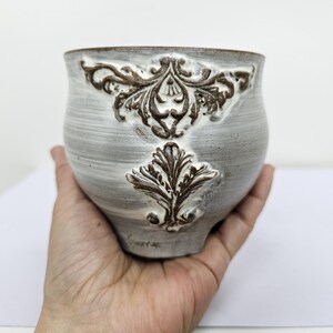 Op de afbeelding: Een grijze keramische kom met een getextureerde afwerking en bruine, in reliëf aangebrachte bloemmotieven. De kom wordt in een hand gehouden, waarbij de afgeronde vorm en decoratieve details worden getoond. Het ontwerp van de kom voegt een vleugje elegantie toe.