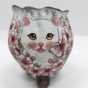 May include: A ceramic planter with a whimsical cat face design. The white planter features a smiling cat face with pink accents, surrounded by pink floral and butterfly details. The planter has three small feet.